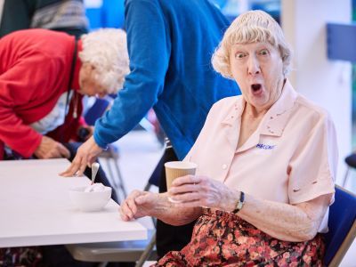 An older lady sits with a cup of tea, pulling a funny face